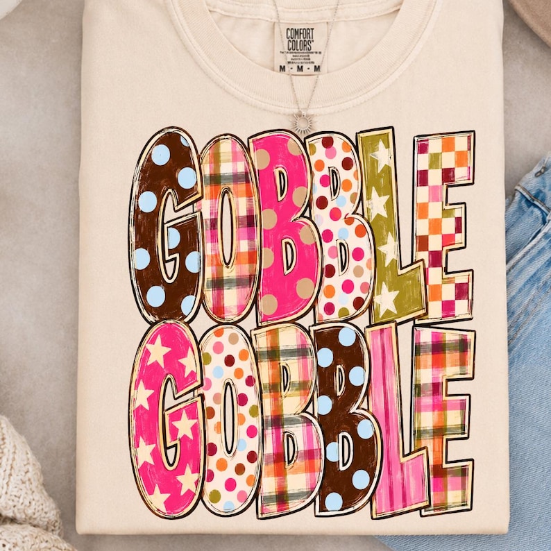 May include: A cream-colored t-shirt featuring the text GOBBLE GOBBLE in two rows. Each letter is filled with a different pattern, including brown with blue dots, plaid, solid pink, multi-colored polka dots, olive green with stars, and orange checks.