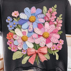 May include: Dark gray t-shirt featuring a vibrant floral design. The design includes large, textured flowers in shades of blue, pink, and white, with a coral-colored bow. The flowers have a patchwork pattern and yellow centers.