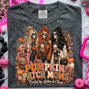 May include: Dark gray Comfort Colors t-shirt featuring a graphic of three skeleton women holding coffee cups, with the text "Pumpkin Patch Moms" and "Fueled by Lattes & Chaos". The design includes pumpkins and fall foliage.
