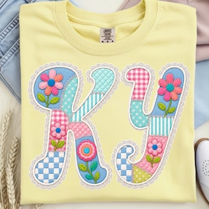 May include: A light yellow t-shirt featuring the letters "KJ" in a patchwork design. Each letter is composed of various patterned fabrics, including polka dots, stripes, and gingham, with floral accents and a white lace trim.