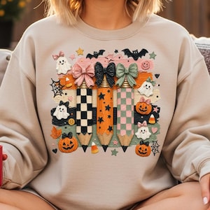 K&ouml;nnte beinhalten: Beiges Sweatshirt mit einem Halloween-Motiv. Das Design zeigt drei Bleistifte mit Schleifen, Sternen und Schachbrettmustern, zusammen mit K&uuml;rbissen, Geistern und Flederm&auml;usen. Das Design ist skurril und festlich.