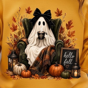 Puede incluir: Sudadera amarilla mostaza con un fantasma con un lazo negro, sosteniendo una taza de café, sentado en un sillón verde. La imagen incluye calabazas, hojas de otoño, una manta a cuadros y un letrero que dice "hello fall".