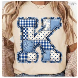 May include: A cream-colored t-shirt featuring a large, patchwork letter "K". The letter is composed of various blue and white patterns, including gingham, polka dots, and a basketball graphic. The edges of the letter are fringed.