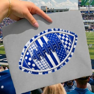 May include: A gray fabric surface featuring a central football-shaped applique. The football is constructed from various blue and white patterned fabric patches, including polka dots, stripes, and gingham, with a frayed edge and white bead trim.