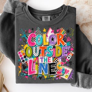 May include: A charcoal gray crewneck sweatshirt featuring a colorful graphic print with the text "COLOR OUTSIDE THE LINES". The design includes illustrations of paint palettes, crayons, paint tubes, and various artistic shapes in vibrant hues.