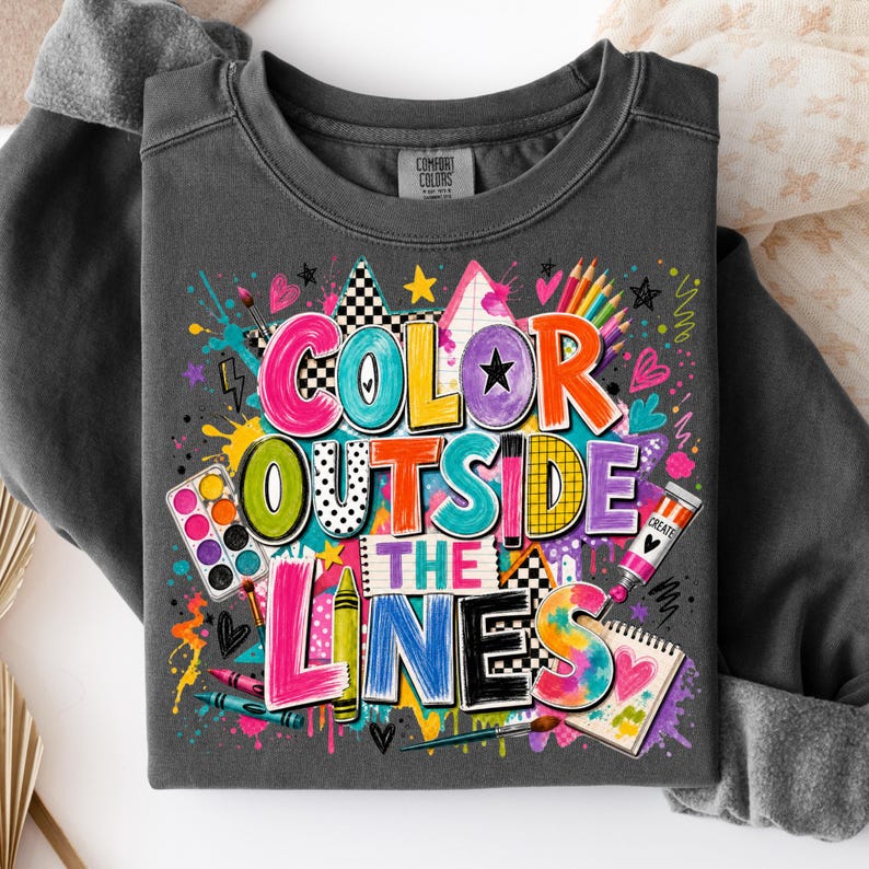May include: A charcoal gray crewneck sweatshirt featuring a colorful graphic print with the text "COLOR OUTSIDE THE LINES". The design includes illustrations of paint palettes, crayons, paint tubes, and various artistic shapes in vibrant hues.