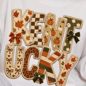 May include: A white sweatshirt featuring the word KENTUCKY in large, textured, embroidered letters. Each letter displays a unique pattern including autumn leaves, brown and white checkers, pumpkins, polka dots, bows, leopard print, and stripes.