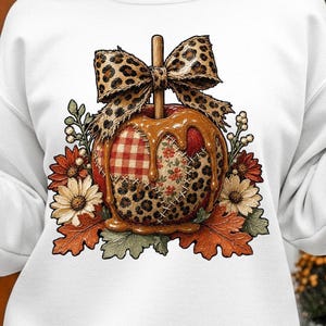 May include: A white long-sleeve sweatshirt featuring a central graphic of a caramel apple with a leopard print bow. The apple has a patchwork design of red gingham, floral, and leopard patterns, surrounded by autumn leaves and flowers.