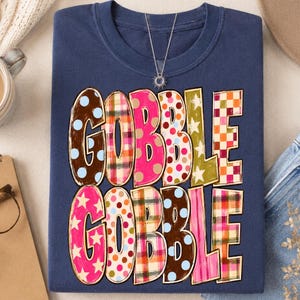 May include: A navy blue crewneck sweatshirt featuring the text GOBBLE GOBBLE in large, blocky letters. Each letter displays a different pattern, including brown with blue dots, pink plaid, solid hot pink, white with orange dots, and green with stars.