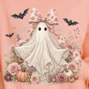 Pode incluir: Uma t-shirt cor de p&ecirc;ssego com o gr&aacute;fico de um fantasma drapeado em renda branca, usando um grande la&ccedil;o com padr&atilde;o floral. O fantasma est&aacute; rodeado por rosas cor-de-rosa, pequenas ab&oacute;boras, teias de aranha e tr&ecirc;s pequenos morcegos.