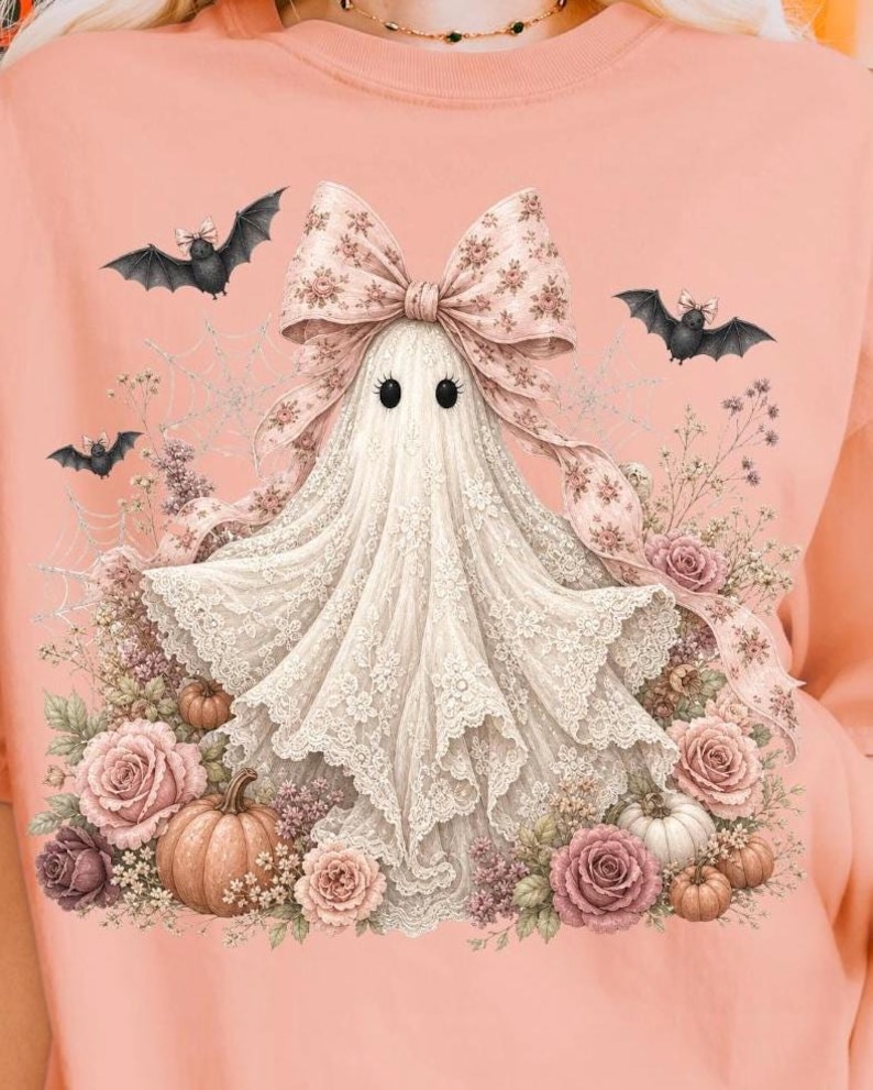 Pode incluir: Uma t-shirt cor de p&ecirc;ssego com o gr&aacute;fico de um fantasma drapeado em renda branca, usando um grande la&ccedil;o com padr&atilde;o floral. O fantasma est&aacute; rodeado por rosas cor-de-rosa, pequenas ab&oacute;boras, teias de aranha e tr&ecirc;s pequenos morcegos.