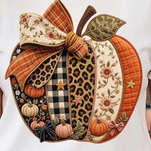 May include: A white t-shirt featuring a large, central pumpkin graphic composed of various fabric patches. The pumpkin design includes sections of leopard print, black and white buffalo plaid, floral patterns, and quilted orange fabric, accented with a fabric bow.