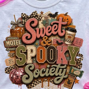 Könnte beinhalten: Weißes T-Shirt mit einem farbenfrohen Grafikdesign mit den Worten "Sweet & Spooky Society". Das Design enthält Kürbisse, Fledermäuse und andere Halloween-Elemente. Das Shirt hat auch ein "Witch's Motel"-Schild und einen Karamellapfel.