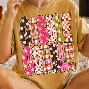 May include: A mustard yellow t-shirt featuring the text GOBBLE GOBBLE in large, bold letters. Each letter is filled with a different colorful pattern, including polka dots, plaid, and stars in shades of pink, brown, orange, green, and blue.