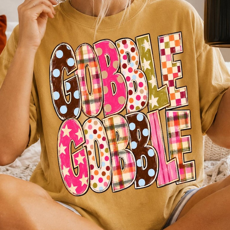 May include: A mustard yellow t-shirt featuring the text GOBBLE GOBBLE in large, bold letters. Each letter is filled with a different colorful pattern, including polka dots, plaid, and stars in shades of pink, brown, orange, green, and blue.