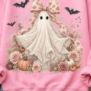 Pode incluir: Uma camisola rosa claro com um gr&aacute;fico central de um fantasma de renda branca usando um grande la&ccedil;o com padr&atilde;o floral. O fantasma est&aacute; rodeado por rosas cor-de-rosa, pequenas ab&oacute;boras e teias de aranha, com dois pequenos morcegos pretos a voar por cima.