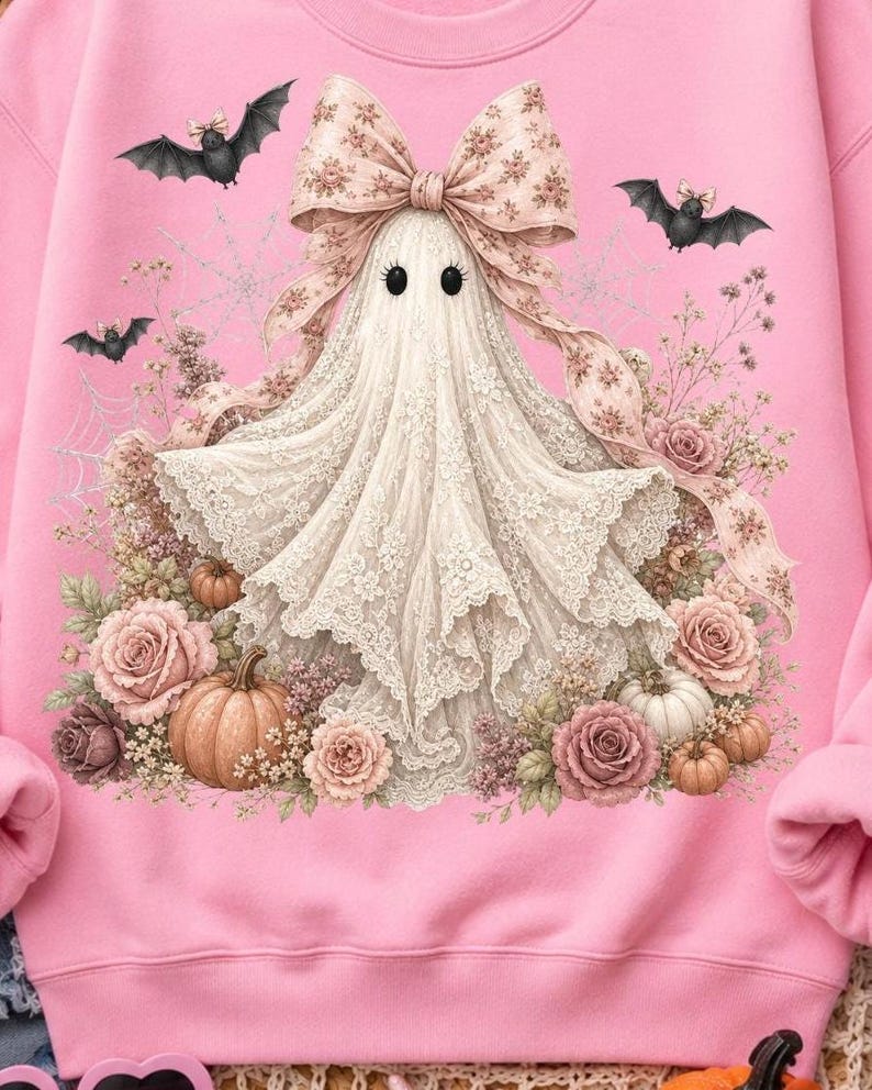 Pode incluir: Uma camisola rosa claro com um gr&aacute;fico central de um fantasma de renda branca usando um grande la&ccedil;o com padr&atilde;o floral. O fantasma est&aacute; rodeado por rosas cor-de-rosa, pequenas ab&oacute;boras e teias de aranha, com dois pequenos morcegos pretos a voar por cima.