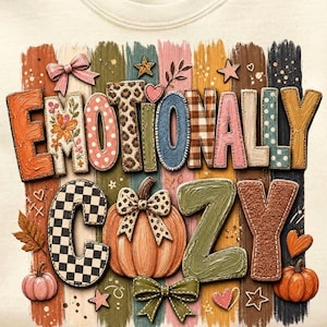 May include: White sweatshirt with the words "Emotionally Cozy" in a colorful, textured design. The design includes pumpkins, bows, stars, and hearts, with a variety of patterns and colors, evoking a fall or autumn theme.