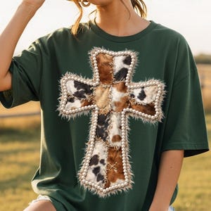 May include: Dark green t-shirt featuring a cross design. The cross is made of patches of cowhide in shades of brown, black, and white, outlined with faux pearls and a fluffy trim. The shirt has short sleeves and a relaxed fit.