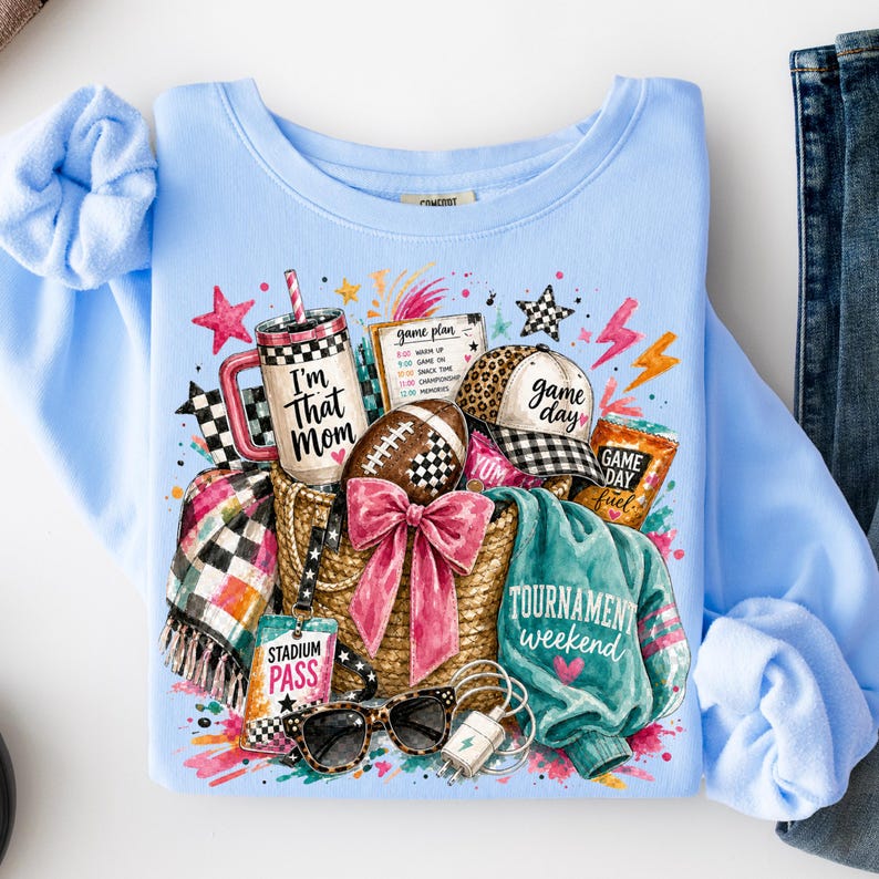 May include: A light blue crewneck sweatshirt featuring a graphic print of a straw tote bag filled with a tumbler labeled I'm That Mom, a football, a leopard print cap, a stadium pass, sunglasses, a plaid blanket, and a teal sweatshirt.