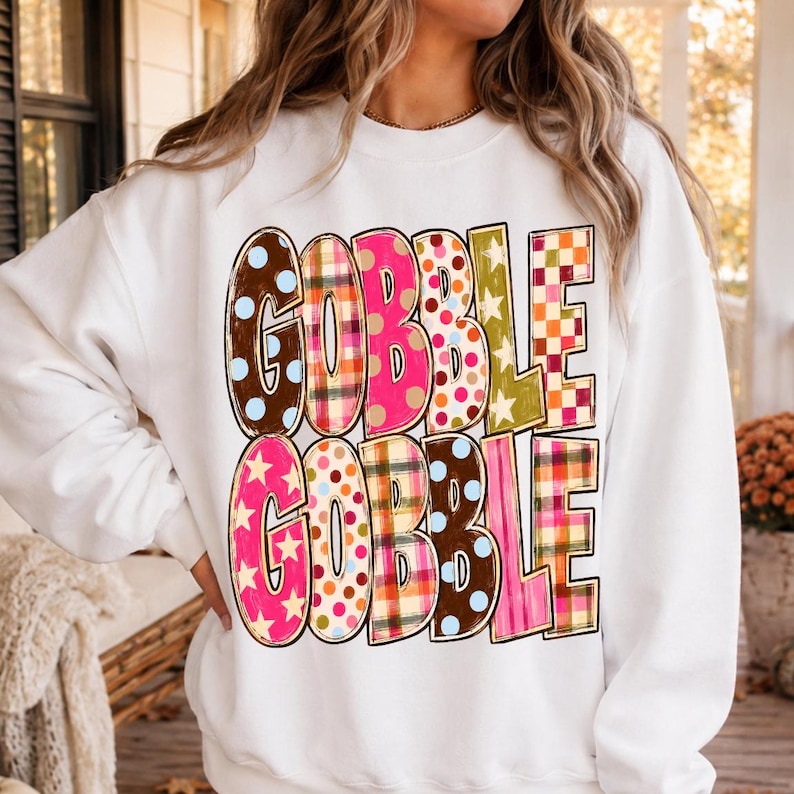 May include: A white long-sleeve sweatshirt featuring the text GOBBLE GOBBLE printed in large, blocky letters. Each letter displays a unique pattern, including polka dots, plaid, and stars in various colors like brown, pink, orange, and green.