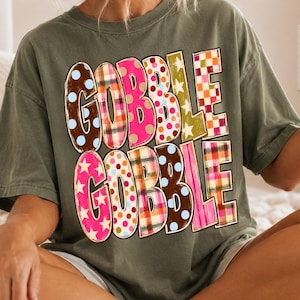 May include: An olive green short-sleeve t-shirt featuring the text GOBBLE GOBBLE in large, blocky letters. Each letter displays a different pattern, including brown with blue dots, pink plaid, solid hot pink, white with pink dots, and orange checkered.