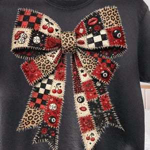 Könnte beinhalten: Dunkelgraues T-Shirt mit einem großen Schleifendesign. Die Schleife ist ein Patchwork aus Mustern, darunter Leopardenmuster, rot-schwarz kariert. Verzierungen sind Kirschen, Würfel, Rosen, Lippen und eine 8-Ball.