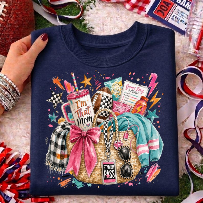 May include: A navy blue crewneck sweatshirt featuring a graphic print of a straw tote bag filled with a tumbler labeled 'I'm That Mom', a football, a game day schedule, a pink bow, sunglasses, and a teal sweater with a plaid scarf.