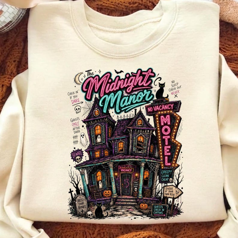 May include: Cream-colored sweatshirt featuring a spooky illustration of "The Midnight Manor." The design includes a haunted house, a "MOTEL" sign, pumpkins, and the text "Check in if you dare." A black cat sits at the bottom.