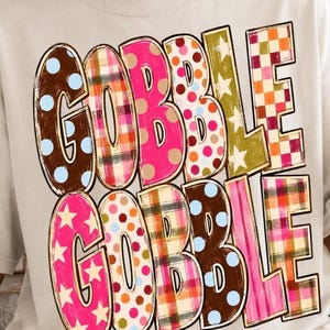May include: A light-colored t-shirt featuring the text GOBBLE GOBBLE in two rows. Each letter is filled with different patterns including brown with blue dots, plaid, pink with dots, olive green with stars, and a red and white checkered pattern.