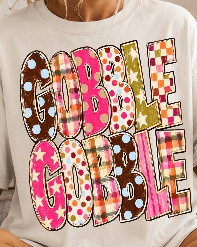 May include: A light-colored t-shirt featuring the text GOBBLE GOBBLE in two rows. Each letter is filled with different patterns including brown with blue dots, plaid, pink with dots, olive green with stars, and a red and white checkered pattern.