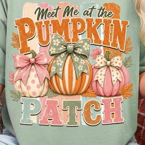 Puede incluir: Camiseta verde claro con un diseño temático de otoño. El diseño incluye tres calabazas con lazos, el texto "Meet Me at the Pumpkin Patch" y hojas de otoño. Las calabazas son en tonos naranja, rosa y crema.