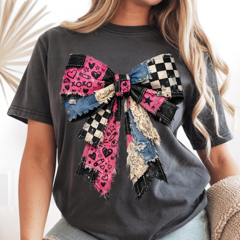 Op de afbeelding: Donkergrijs t-shirt met een groot strikdesign. De strik is een patchwork van denim, roze stof met hartjes en XOXO, zwart-wit geblokt en kant. De strik heeft een versleten, vintage uitstraling.