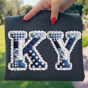May include: A dark gray fabric item with the letters "KY" appliqued on the front. The letters are made of patchwork fabric in blue and white patterns, including gingham, floral, and polka dots, with a white lace trim.