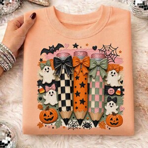 K&ouml;nnte beinhalten: Pfirsichfarbenes Sweatshirt mit einem Halloween-Motiv. Das Design umfasst drei bleistiftf&ouml;rmige Figuren mit Schleifen, Sternen und Karomustern. Geister, K&uuml;rbisse und Spinnweben vervollst&auml;ndigen den festlichen Look. Ein lustiges und einzigartiges Oberteil f&uuml;r die gruselige Jahreszeit.
