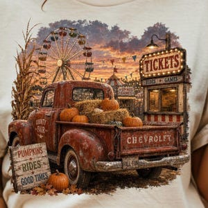 Op de afbeelding: T-shirt met een vintage rode Chevrolet-truck vol pompoenen en hooibalen, tegen een kermisachtergrond met een reuzenrad en een ticketkraam. Het shirt heeft de woorden "County Fair", "Pumpkins Rides Good Times" en "Fall-Fun".