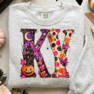Pode incluir: Uma camisola cinzenta clara de gola redonda com um grande estampado gr&aacute;fico das letras KV. O K est&aacute; preenchido com um c&eacute;u noturno roxo, uma lua crescente, uma casa assombrada e uma ab&oacute;bora. O V apresenta bolas cor de laranja e pretas.