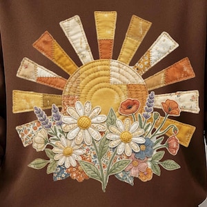 Könnte beinhalten: Ein braunes Sweatshirt mit einer großen, gesteppten Sonnenapplikation in Gelb-, Orange- und Cremetönen. Unter der Sonne befindet sich ein detailliertes Blumenarrangement mit weißen Gänseblümchen, orangefarbenen Mohnblumen und Lavendelzweigen aus Stoff.