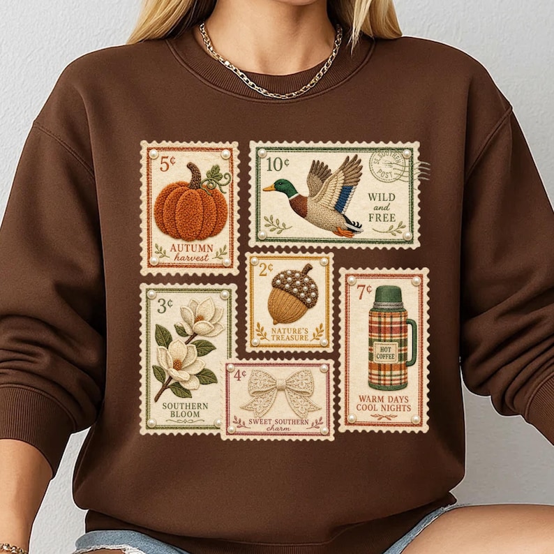 May include: A brown sweatshirt featuring postage stamp designs with autumn and Southern themes. Stamps depict a pumpkin, duck, acorn, bow, magnolia, and a thermos. Text includes "Autumn Harvest," "Wild and Free," "Nature's Treasure," "Southern Bloom," "Sweet Southern Charm," and "Warm Days Cool Nights."