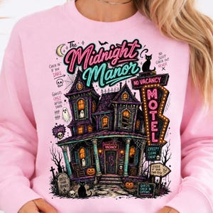 May include: Pink sweatshirt featuring a detailed illustration of a haunted house with the text "The Midnight Manor." The design includes a motel sign, pumpkins, ghosts, and the words "Vacancy" and "Beware."