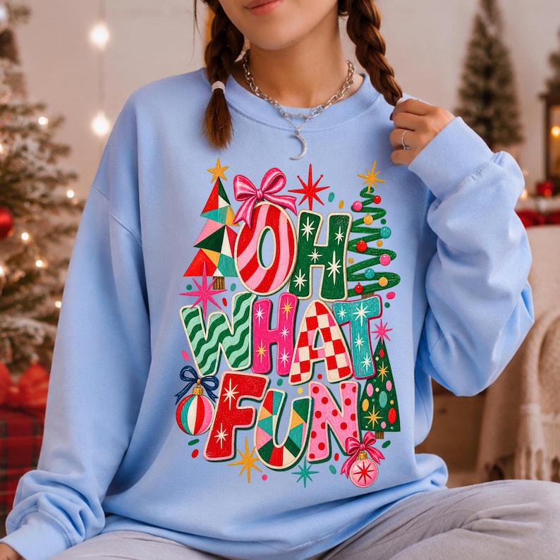 K&ouml;nnte beinhalten: Ein hellblaues Sweatshirt mit Rundhalsausschnitt und einem bunten Grafikdruck mit dem Text "OH WHAT FUN". Das Design zeigt stilisierte Weihnachtsb&auml;ume, Ornamente, Sterne und eine rosa Schleife in verschiedenen Mustern wie Streifen, Karos und Punkten.