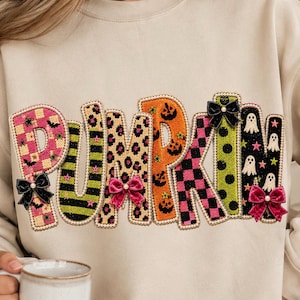 May include: A beige sweatshirt featuring the word PUMPKIN in large, textured letters. Each letter has a unique pattern including pink and orange checkers, leopard print, stripes, and ghosts. The design is accented with small black and pink bows.