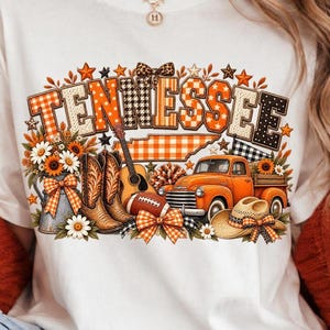Może przedstawiać: Biały t-shirt z nadrukiem graficznym przedstawiającym słowo TENNESSEE w pomarańczowe i czarne wzory. Projekt zawiera mapę stanu, pomarańczowy pickup, brązowe kowbojki, gitarę akustyczną, piłkę do futbolu amerykańskiego oraz słoneczniki.
