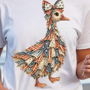 Può includere: T-shirt bianca con una dettagliata illustrazione di un'anatra decorata con tessuto rosso, bianco e blu, che ricorda la bandiera americana. L'anatra ha un fiocco e sta camminando. Il design è colorato e patriottico.