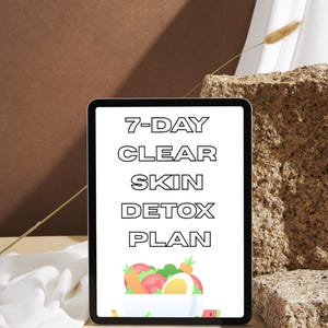 May include: A digital tablet displays a "7-DAY CLEAR SKIN DETOX PLAN" in bold black text. Below the text is a colorful illustration of a bowl filled with fruits and vegetables. The tablet is set against a neutral background with decorative elements.
