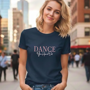 Könnte beinhalten: Ein marineblaues T-Shirt mit dem Text "DANCE Your Heart Out" in rosa Schrift. Das Shirt hat einen Rundhalsausschnitt und wird von einer Person im Freien getragen.