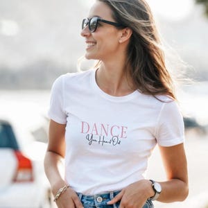 Könnte beinhalten: Weißes T-Shirt mit dem Text "DANCE Your Heart Out" in rosa und schwarzer Schrift. Das T-Shirt hat einen Rundhalsausschnitt. Die Person, die das T-Shirt trägt, trägt auch eine Sonnenbrille und eine Uhr.