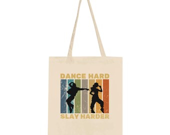 May include: Cream-colored tote bag with the text "DANCE HARD SLAY HARDER" in a retro font. The design features black silhouettes of two dancers against a backdrop of vertical stripes in various colors. The bag has long, cream-colored handles.
