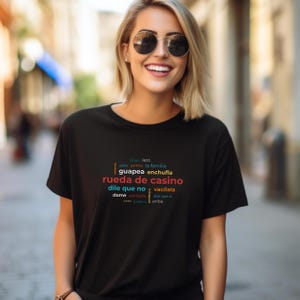 May include: A black t-shirt with colorful text in Spanish, including "rueda de casino" in red. The shirt is worn by a person with blonde hair and sunglasses, smiling in an outdoor setting.