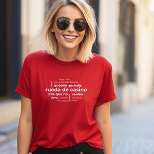 May include: A red t-shirt with white text. The text includes phrases like "rueda de casino" and "dile que no." The shirt is a crew neck style. The person in the image is wearing sunglasses.